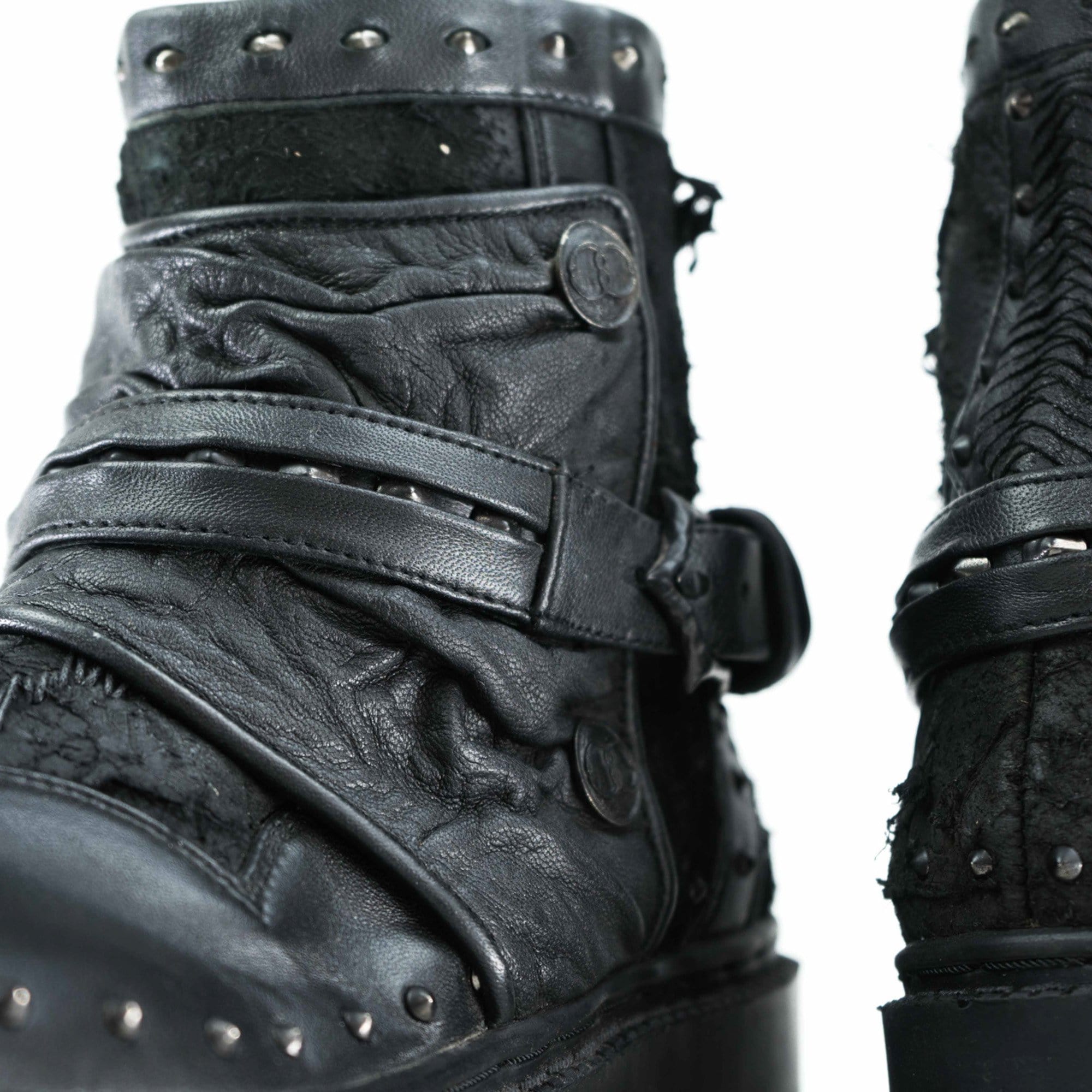 Close-up of a pair of worn, black leather boots with a rugged design. These MAGMA Wasteland Studded Leather Ankle Combat Boots from Kaya Movement feature decorative metal studs, textured leather, straps, and buckles. The material appears distressed, giving the studded leather ankle boots a unique, vintage look. The background is white.
